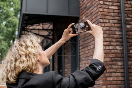 Blonde Immobilienmaklerin mit Handy fotografiert modernes Haus an der Stadtstraße