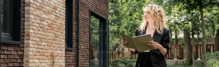 attractive blonde real estate agent with folder looking at building on urban street, banner