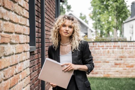 Photo for Blonde and stylish property realtor holding folder and looking at camera near building on street - Royalty Free Image