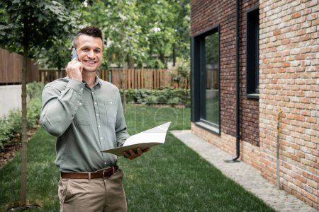 fröhlicher Immobilienmakler mit Ordner, der auf dem Smartphone spricht und in die Kamera in der Nähe des Hauses blickt