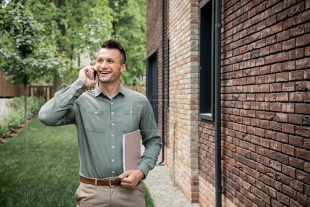 pleased real estate agent with folder looking away while talking on mobile phone near cottage