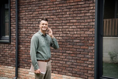 erfolgreicher Immobilienmakler hält Ordner in der Hand und telefoniert mit dem Handy in der Nähe eines Gebäudes auf der Straße