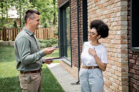 Immobilienmakler mit Mappe und Stift im Gespräch mit lächelndem afrikanisch-amerikanischem Kunden in der Nähe von neuem Ferienhaus