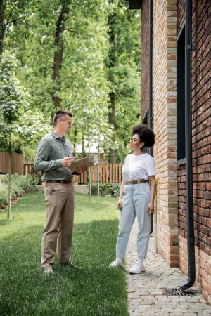 Téléchargez les photos : Pleine longueur de confiant agent immobilier parler à une femme afro-américaine près de bâtiment dans la rue - en image libre de droit