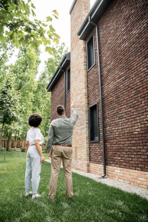 vue arrière du courtier immobilier pointant avec la main et montrant une maison neuve à un client afro-américain