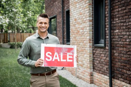 happy and confident real estate agent looking at camera and holding sale signboard near new house