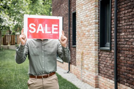 Photo for Real estate agent obscuring face with sale signboard while standing near cottage in city - Royalty Free Image