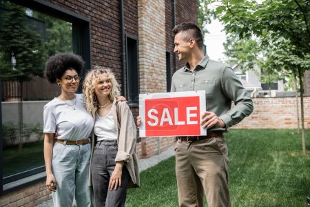 Photo for Real estate agent holding sale signboard near cheerful interracial lesbian women and new cottage - Royalty Free Image