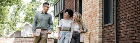 joyful multicultural lesbian couple talking with real estate agent near house on street, banner