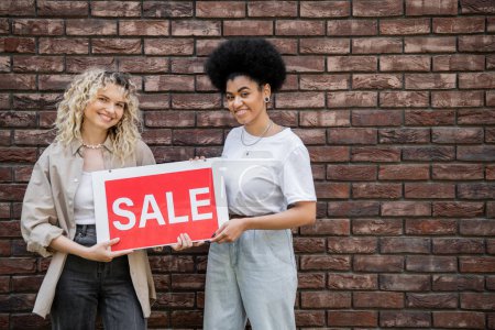 cheerful multiethnic lesbian couple holding sale signboard and looking at camera near cottage