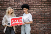 cheerful multiethnic lesbian couple holding sale signboard and looking at camera near cottage tote bag #664925286