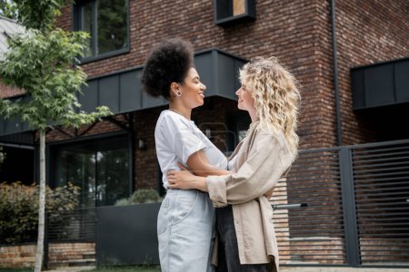 Photo for Side view of interracial lesbian couple embracing and smiling at each other near new own cottage - Royalty Free Image