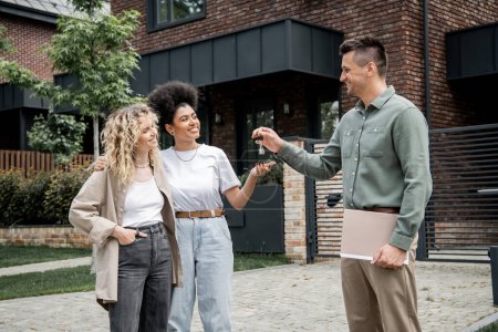 successful realtor with folder giving key from new house to pleased multiethnic lesbian couple
