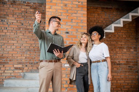 real estate agent pointing with hand near smiling interracial lesbian women in new cottage
