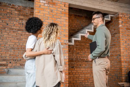 realtor pointing at stairs and showing house with unfinished interior to lesbian interracial couple