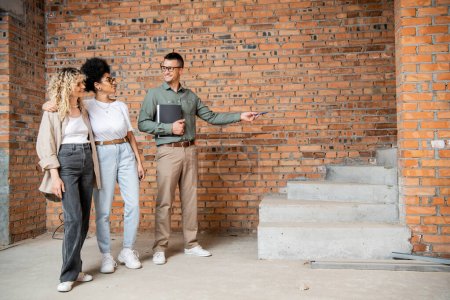 Lächelnder Immobilienmakler zeigt auf Treppen, während er lesbischem multiethnischen Paar sein neues Haus zeigt