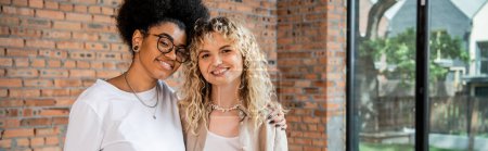 pleased interracial lesbian couple embracing and smiling at camera in new house, banner