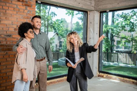 Lächelnder Immobilienmakler zeigt mit der Hand auf neue Wohnung für multiethnisches Paar