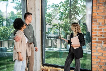 real estate agent with folder pointing with hand at window in new cottage near interracial couple