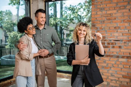joyful real estate agent with folder holding keys near happy multicultural couple in new cottage