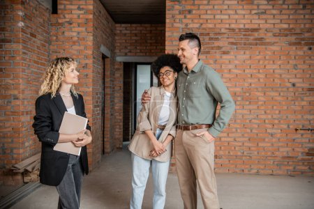 pleased multiethnic couple embracing near real estate agent standing with folder in new cottage