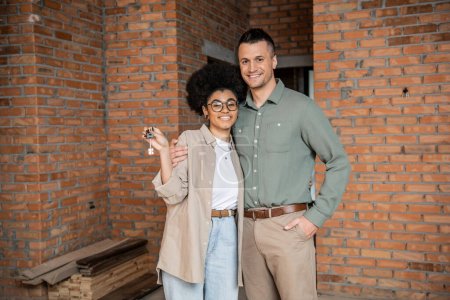 overjoyed interracial couple with keys looking at camera in new private cottage