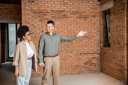 glücklicher Mann zeigt mit der Hand, während er der afrikanisch-amerikanischen Frau ein neues Haus zeigt