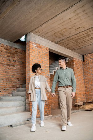 Photo for Joyful interracial couple holding hands and looking at each other while walking in new cottage - Royalty Free Image