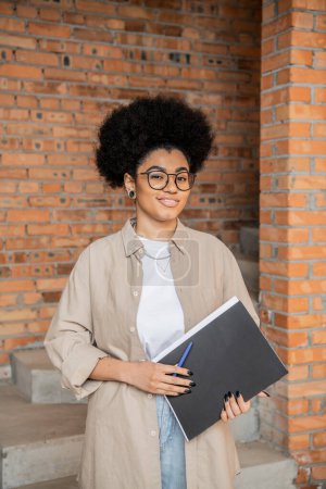 african american real estate agent in eyeglasses holding folder and smiling at camera in new house