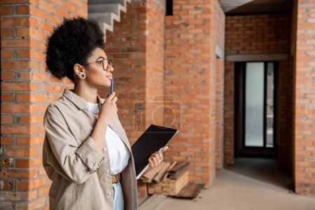 Téléchargez les photos : Vue de côté de réfléchi agent immobilier afro-américain debout avec stylo et dossier dans une maison neuve - en image libre de droit