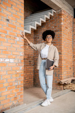 Téléchargez les photos : Toute la longueur de l'agent immobilier afro-américain avec dossier regardant loin dans la maison de ville neuve - en image libre de droit