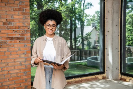 joyeux agent immobilier afro-américain avec dossier regardant la caméra dans la maison avec de grandes fenêtres