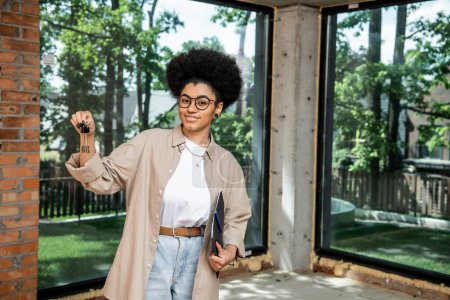 Photo for Positive african american real estate agent holding folder and keys in modern city cottage - Royalty Free Image