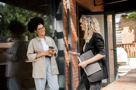 blonde real estate broker talking to african american woman with coffee to go near cottage