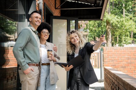 real estate agent pointing with pen near multiethnic couple standing near modern cottage