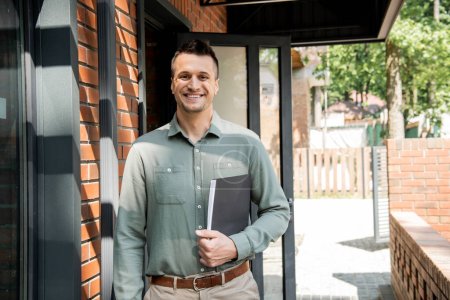 successful and happy real estate agent with folder looking at camera near modern cottage