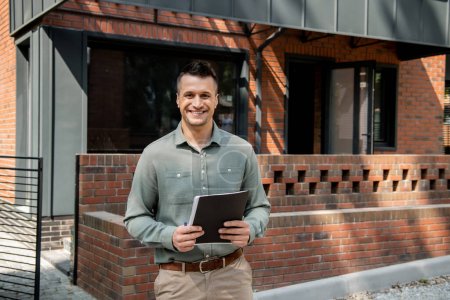 confident real estate agent holding folder and looking at camera near modern house on street