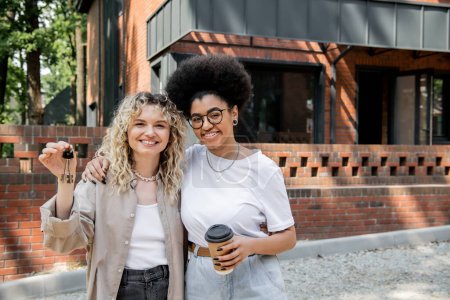 Téléchargez les photos : Gai interracial lesbienne couple avec clés et café pour aller regarder caméra près de maison privée - en image libre de droit
