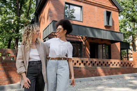 joyful multiethnic lesbian couple looking at each other next to private modern cottage magic mug #664926208