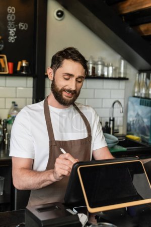 smiling bearded barista in apron using digital tablet while working on bar in coffee shop