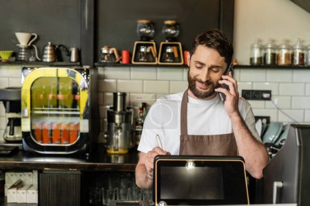 smiling barista in apron talking on smartphone and using digital tablet in coffee shop