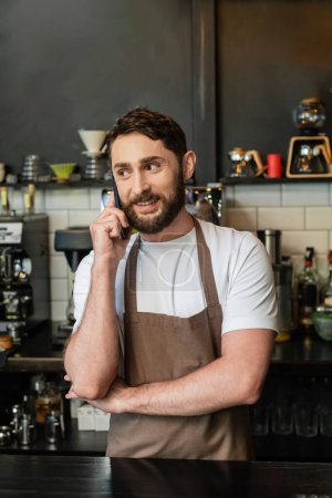 positive bearded barista in apron talking on smartphone while standing near bar in coffee shop