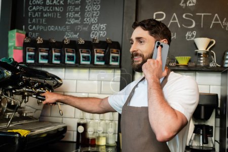 Lächelnder Barista in Schürze, der auf dem Smartphone spricht, während er in der Nähe der Kaffeemaschine im Café arbeitet