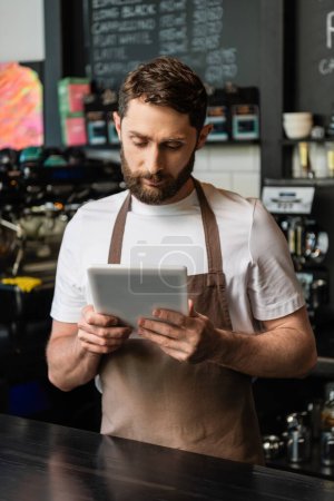 barista barbudo en delantal usando tableta digital mientras trabaja y está de pie cerca del bar en la cafetería