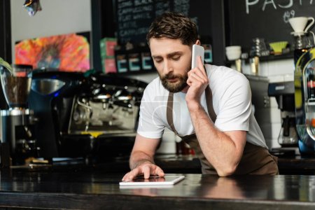 barista in apron talking on smartphone and using digital tablet on bar in coffee shop