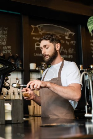 bearded barista in apron cleaning coffee machine holder with brush in coffee shop