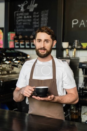 sonriente barbudo barista en delantal sosteniendo taza de café y mirando a la cámara en la cafetería