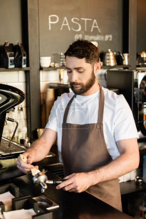 bearded barista in apron cleaning holder with brush near coffee machine while working in cafe