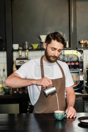 bearded barista in apron pouring milk from pitcher while making cappuccino on bar in coffee shop