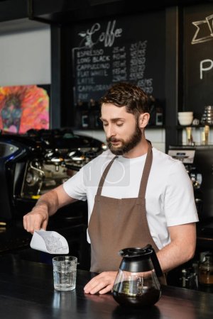 barista in apron pouring ice cubes in glass near coffee pot on bar in coffee shop